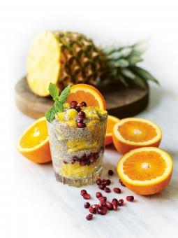 Chiafrögröt i glas med apelsin ananas granatäppelkärnor och en kvist mynta på toppen. Apelsiner och resten av ananasen runt om