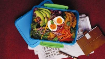 Blå matlåda fylld med ägg, avocado, morot, blandade frön, rödkål spenat mm stående ovanpå sitt lock och ett korsord